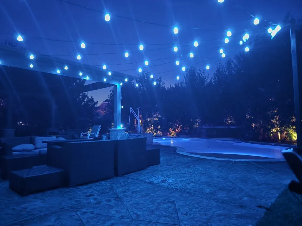 Blue Backyard String Lights and Pool – Houston - Laymance Lighting Color-changing string lights glowing blue over a Houston backyard pool and patio, with landscape uplighting on the trees by Laymance Lighting