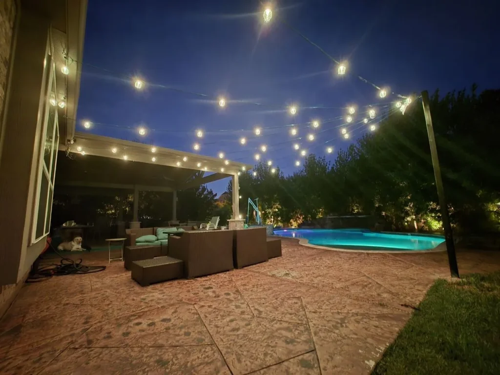 Warm White Patio String Lights and Pool – Houston - Laymance Lighting Warm white string lights crisscrossing over a Houston backyard patio and pool, with landscape uplighting on the trees, installed by Laymance Lighting