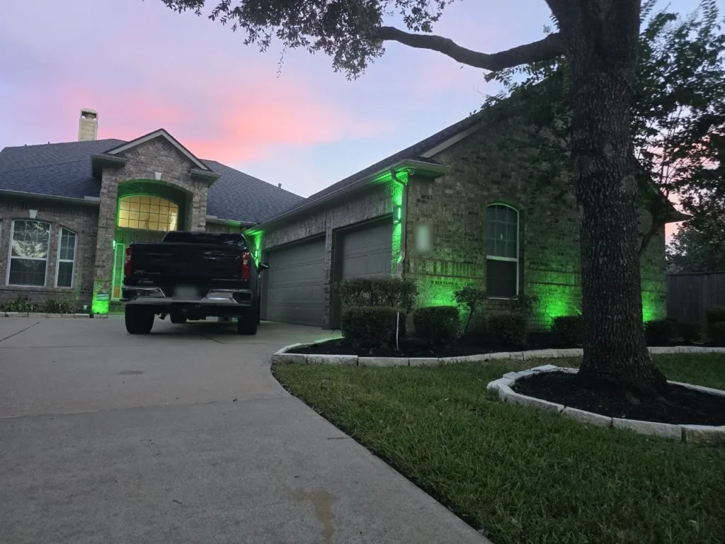 Green Color-Changing Landscape Lighting – Houston - Laymance Lighting Houston brick home at dusk with app-controlled color-changing landscape lighting set to green, washing the walls, entry, and garage by Laymance Lighting