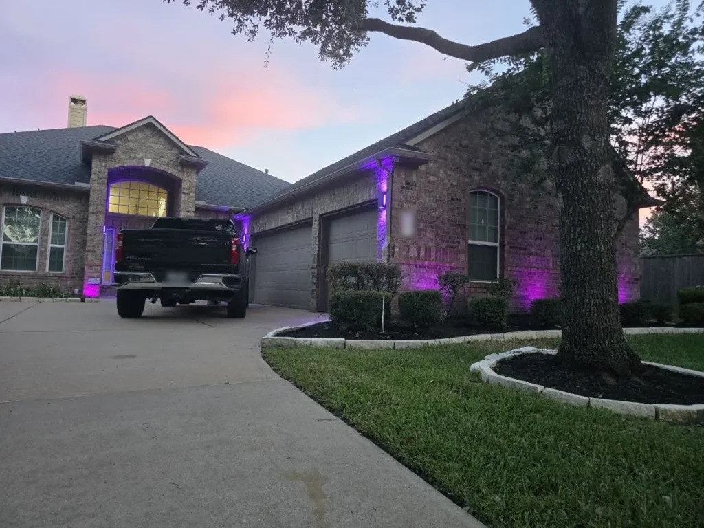 Purple Color-Changing Landscape Lighting – Houston - Laymance Lighting Houston brick home at dusk with color-changing landscape uplights set to purple, highlighting the arched entry and garage, by Laymance Lighting